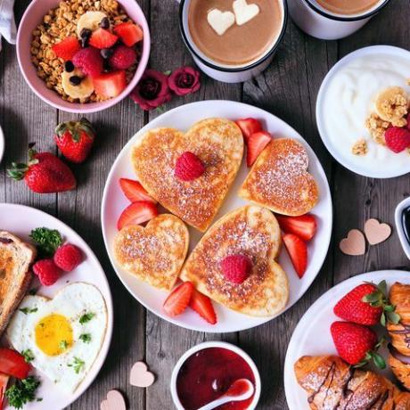 adegan meja sarapan hari kasih sayang atau ibu dengan latar belakang kayu gelap dengan panekuk berbentuk hati, telur, dan makanan bertema cinta